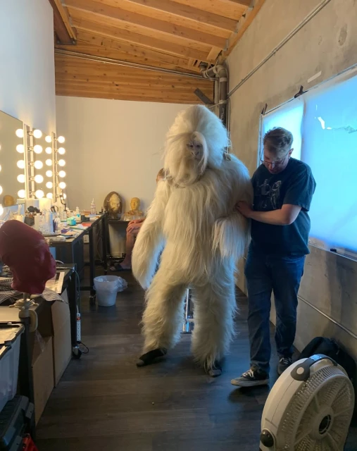 Costumed performer in a white furry suit poses in a backstage dressing room with a makeup table and a crew member adjusting the suit.